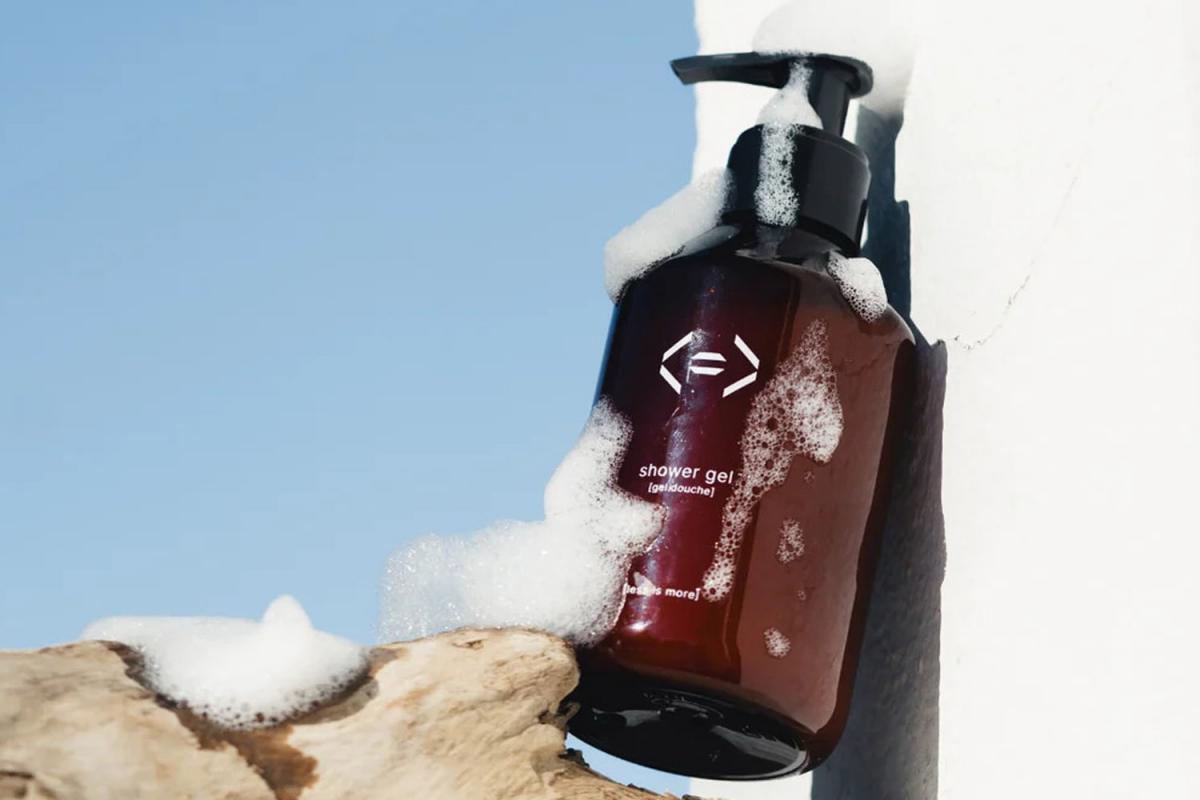 Duschgel mit Pumpspender und Schaum außen an der Flasche lehnt an einer Mauer. Der Untergrund ist Schwemmholz, im Hintergrund ist blauer Himmel zu sehen