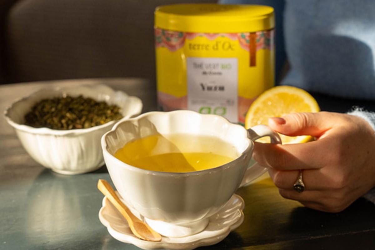 Eine Frauenhand hält eine weiße Tasse mit Grüntee mit Yuzu. Im Hintergrund sieht man eine gelbe Teedose von Terre D'Oc. 