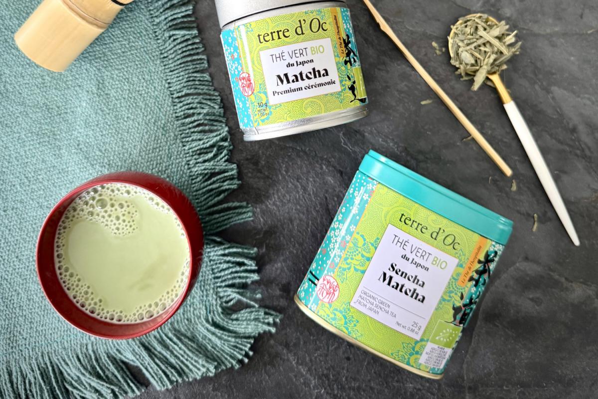 Matcha-Tee in einer Tasse auf einem türkisen Tischtuch. Daneben liegt eine Metalldose mit Matcha-Tee und darüber eine kleinere mit dem selben Inhalt.